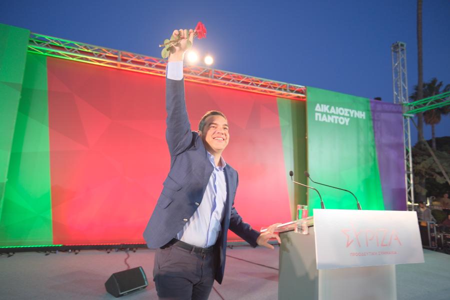 Ο Αλέξης Τσίπρας στη σκηνή με το χέρι σηκωμένο να κρατά ένα γαρύφαλλο 