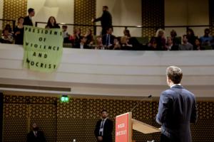 Ο Εμανουελ Μακρόν στο βήμα, γυρισμένος πλάτη κοιτά στο θεωρείο τους διαδηλωτές που κρατούν ένα πανό