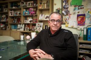 Ο Γιάννης Μουζάλας καθισμένος σε καρέκλα με τα χέρια σταυρωμένα δίπλα σε ένα τραπέζι και πίσω του ράφια με κορνίζες