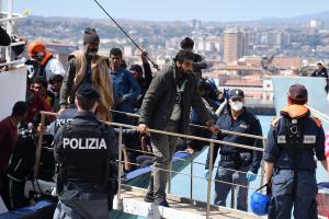 Ένας αστυνομικός της Ιταλίας γυρισμένος πλάτη κοιτά προς το σκάφος από το οποίο βγαίνουν μετανάστες