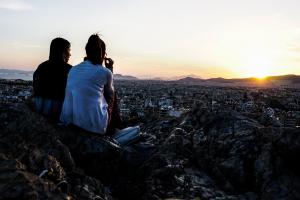 Δύο κορίτσια κάθονται σε βραχάκια και βλέπουν το ηλιοβασίλεμα με θέα την Αθήνα
