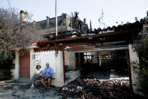 ζευγάρι ηλικιωμένων κάθεται έξω από το κατεστραμμένο από την φωτιά σπίτι του στο Λαγονήσι