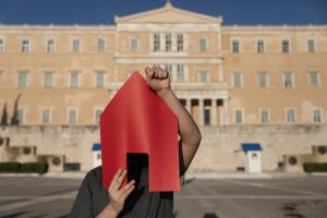άντρας με κόκκινο χαρτόνι σε σχήμα σπιτιού μπροστά στο πρόσωπό του και σηκωμένη την γροθιά του σε διαμαρτυρία μπροστά στη Βουλή