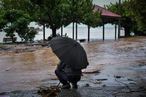 Άντρας με μαύρη ομπρέλα καθισμένος πλάτη σε λασπωμένο δρόμο από τις πλημμύρες