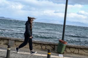 Κοπέλα με μπουφάν περπατά δίπλα στην θάλασσα με τον αέρα να της παίρνει τα μαλλιά