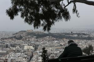 Άντρας καθισμένος σε παγκάκι κοιτά προς την Ακρόπολη και τις πολυκατοικίες στην Αθήνα