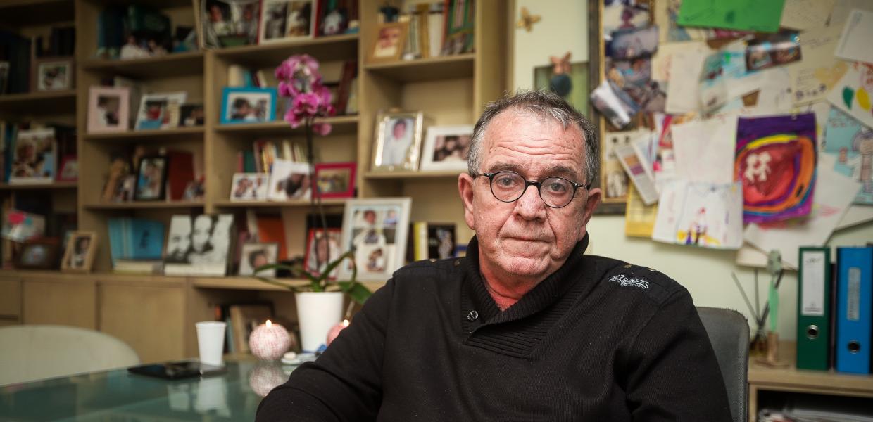 Ο Γιάννης Μουζάλας καθισμένος σε καρέκλα με τα χέρια σταυρωμένα δίπλα σε ένα τραπέζι και πίσω του ράφια με κορνίζες