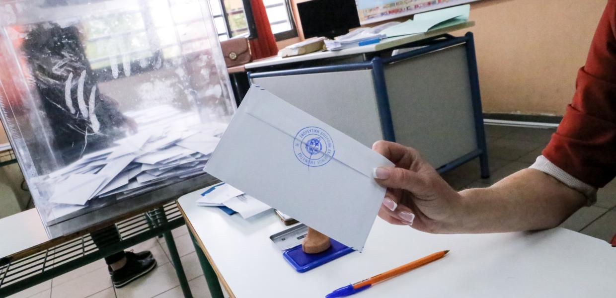 χέρι κρατά φάκελο και πίσω η κάλπη και η έδρα 