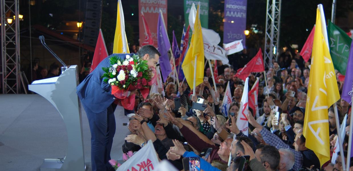 Ο Αλέξης Τσίπρας κρατά λουλούδια και χαιρετά κόσμο που βρίσκεται κάτω από την σκηνή
