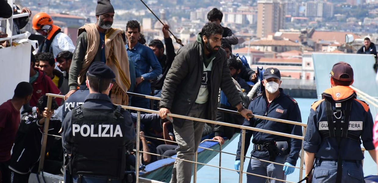 Ένας αστυνομικός της Ιταλίας γυρισμένος πλάτη κοιτά προς το σκάφος από το οποίο βγαίνουν μετανάστες