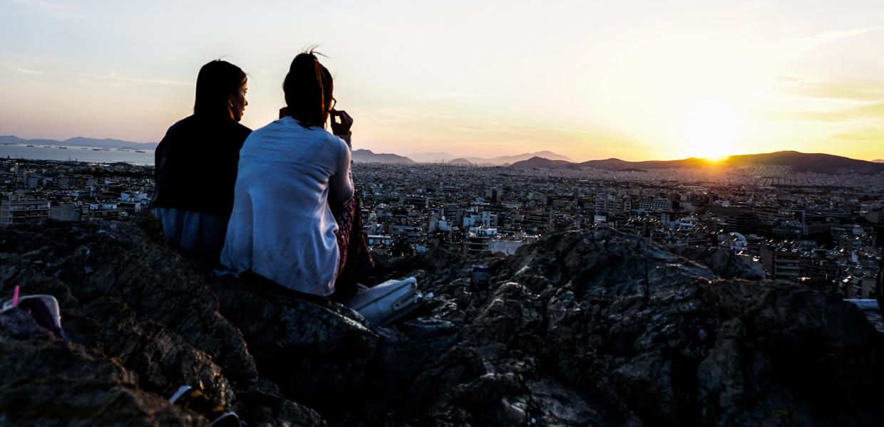 Δύο κορίτσια κάθονται σε βραχάκια και βλέπουν το ηλιοβασίλεμα με θέα την Αθήνα