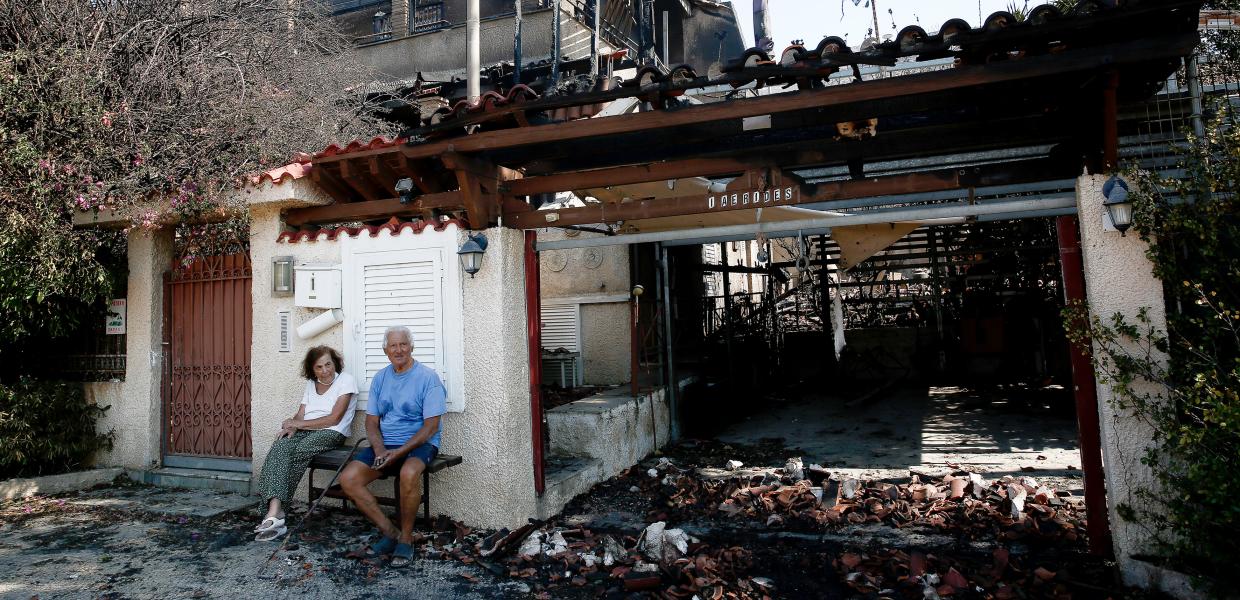 ζευγάρι ηλικιωμένων κάθεται έξω από το κατεστραμμένο από την φωτιά σπίτι του στο Λαγονήσι