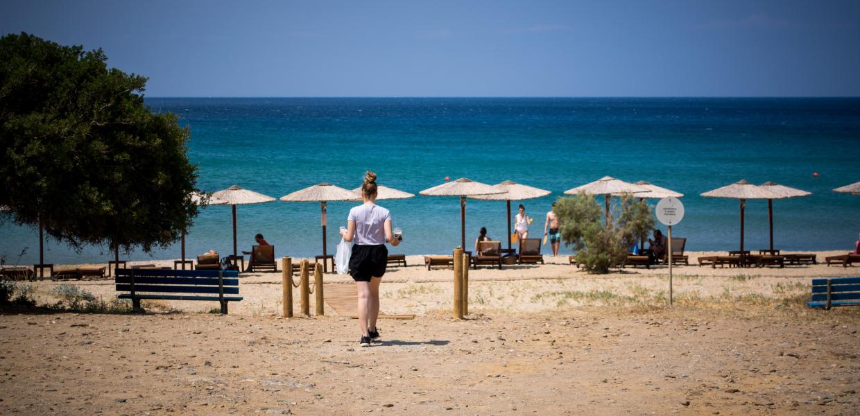 κοπέλα σε σακούλα και καφέ στο χέρι πάει προς θάλασσα με ομπρέλες και ξαπλώστρες