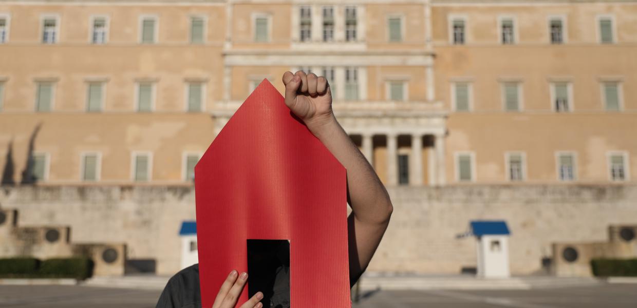 άντρας με κόκκινο χαρτόνι σε σχήμα σπιτιού μπροστά στο πρόσωπό του και σηκωμένη την γροθιά του σε διαμαρτυρία μπροστά στη Βουλή