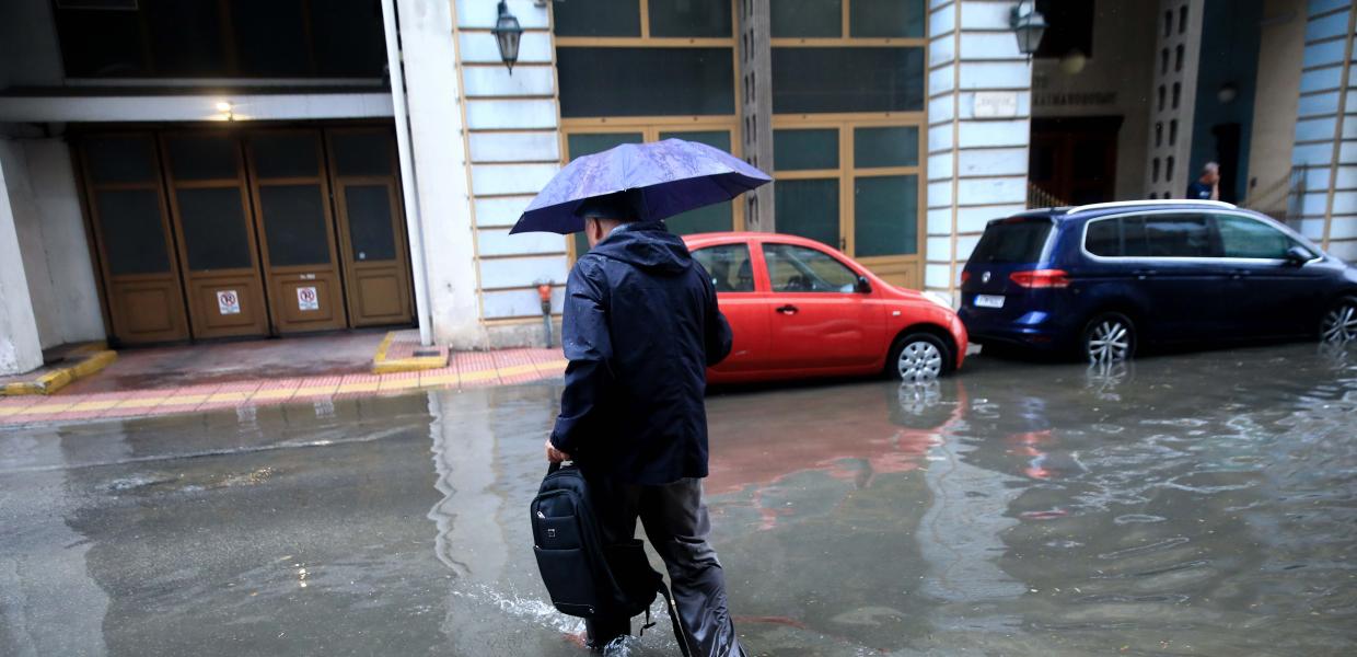 Άντρας με ομπρέλα περπατά σε δρόμο που έχει γεμίσει νερά από την βροχή