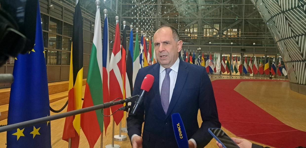 Ο Γεραπετρίτης στις Βρυξέλλες μιλά σε μικρόφωνα δημοσιογράφων και πίσω του οι σημαίες χωρών της ΕΕ