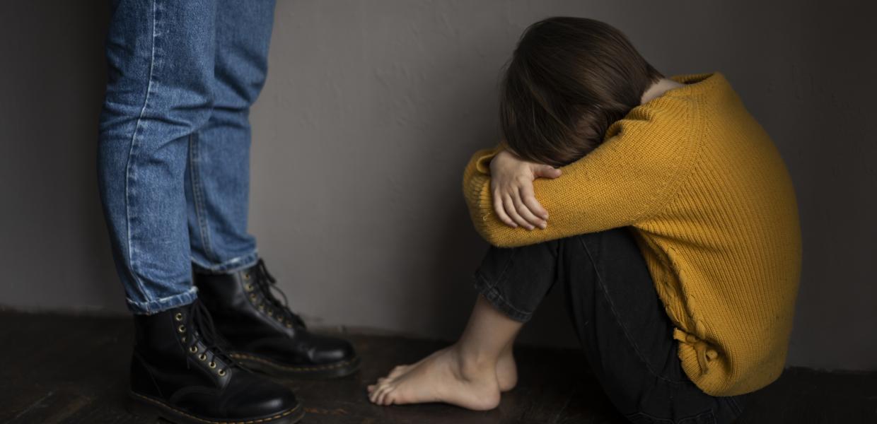 παιδάκι καθισμένο στο πάτωμα με το κεφάλι κρυμμένο στα πόδια και ενήλικας που στέκεται όρθιος μπροστά του