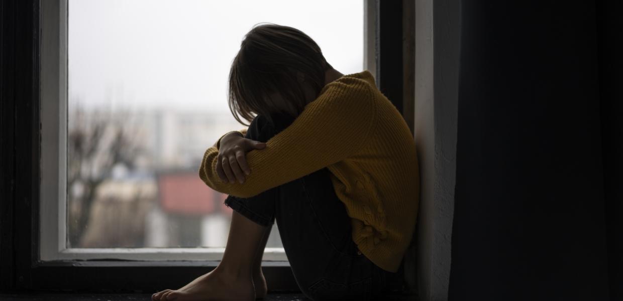 Κοριτσάκι φοβισμένο κάθεται σε περβάζι παραθύρου με το πρόσωπο στα γόνατά της