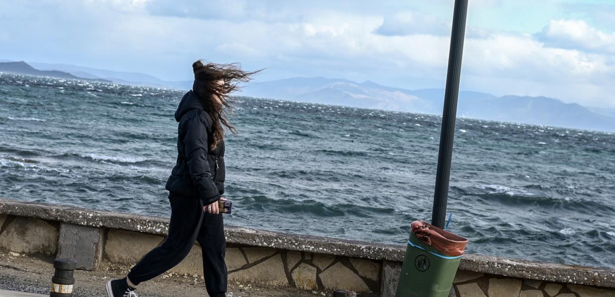 Κοπέλα με μπουφάν περπατά δίπλα στην θάλασσα με τον αέρα να της παίρνει τα μαλλιά