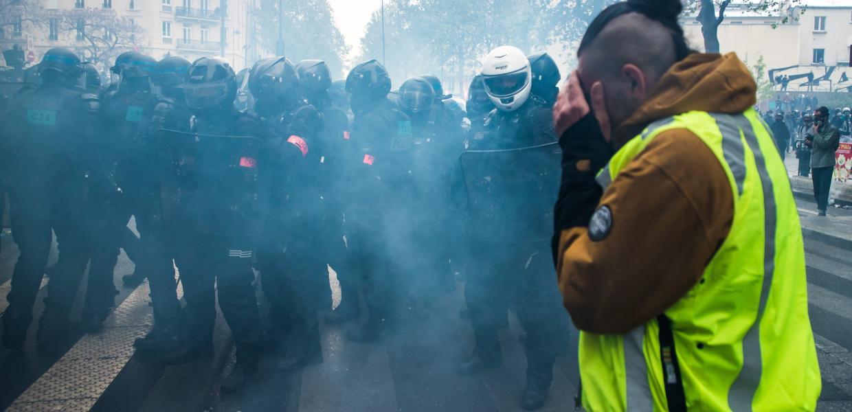 Γάλλος διαδηλωτής με κίτρινο γιλέκο μπροστά από ΜΑΤ προσπαθεί να προστατευτεί από δακρυγόνο